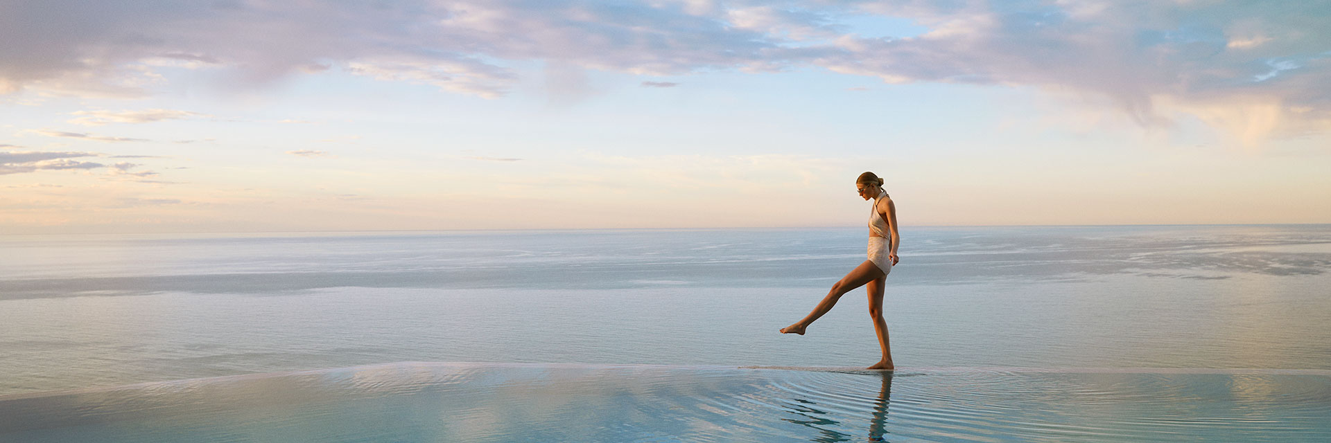 A lady in a swimsuit walks along the infinity pool edge infront of the sea and a beautiful sunset